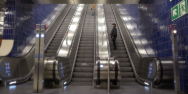 Breite Rolltreppenanlage in einer U-Bahn-Halle mit dunkelblauen Fliesenwänden. Zwei Personen stehen auf verschiedenen Rolltreppen und wenden sich aneinander zu.