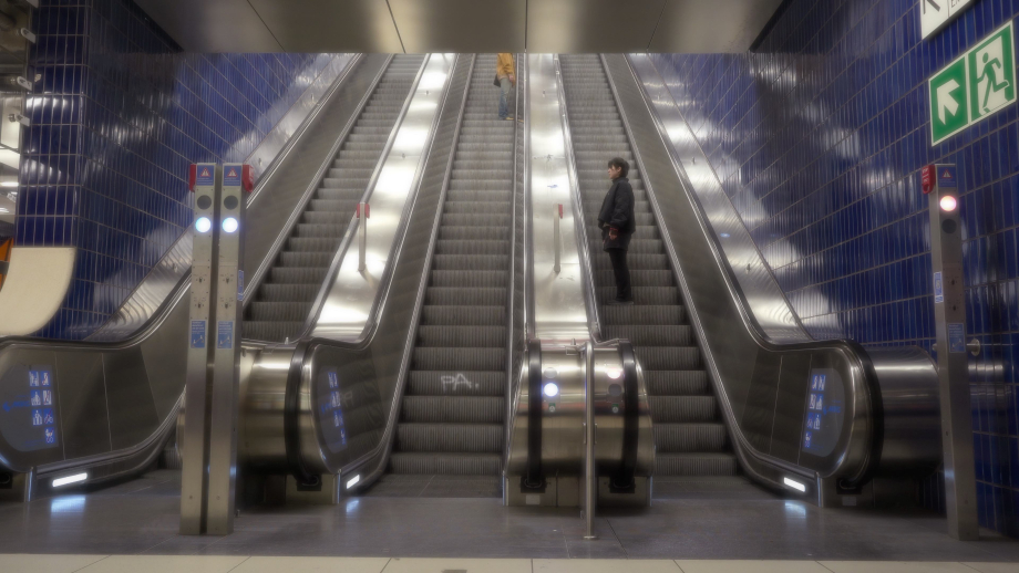 Breite Rolltreppenanlage in einer U-Bahn-Halle mit dunkelblauen Fliesenwänden. Zwei Personen stehen auf verschiedenen Rolltreppen und wenden sich aneinander zu.