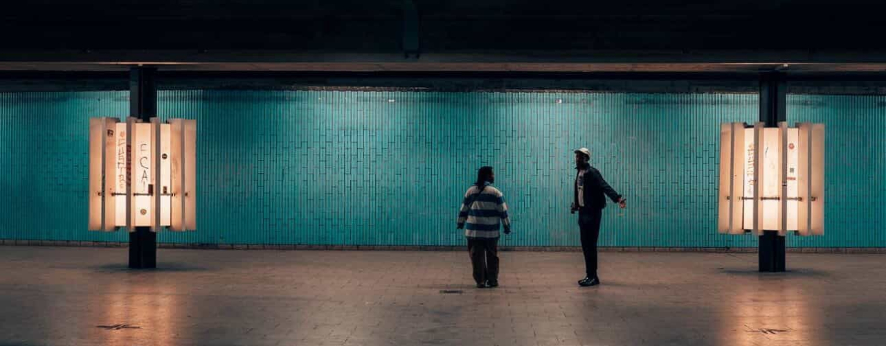 Zwei Personen stehen sich in einem weiten, türkis geflieste Raum gegenüber, der wie eine Unterführung aussieht. Warme Leuchtkästen an den Wänden erhellen den Raum.