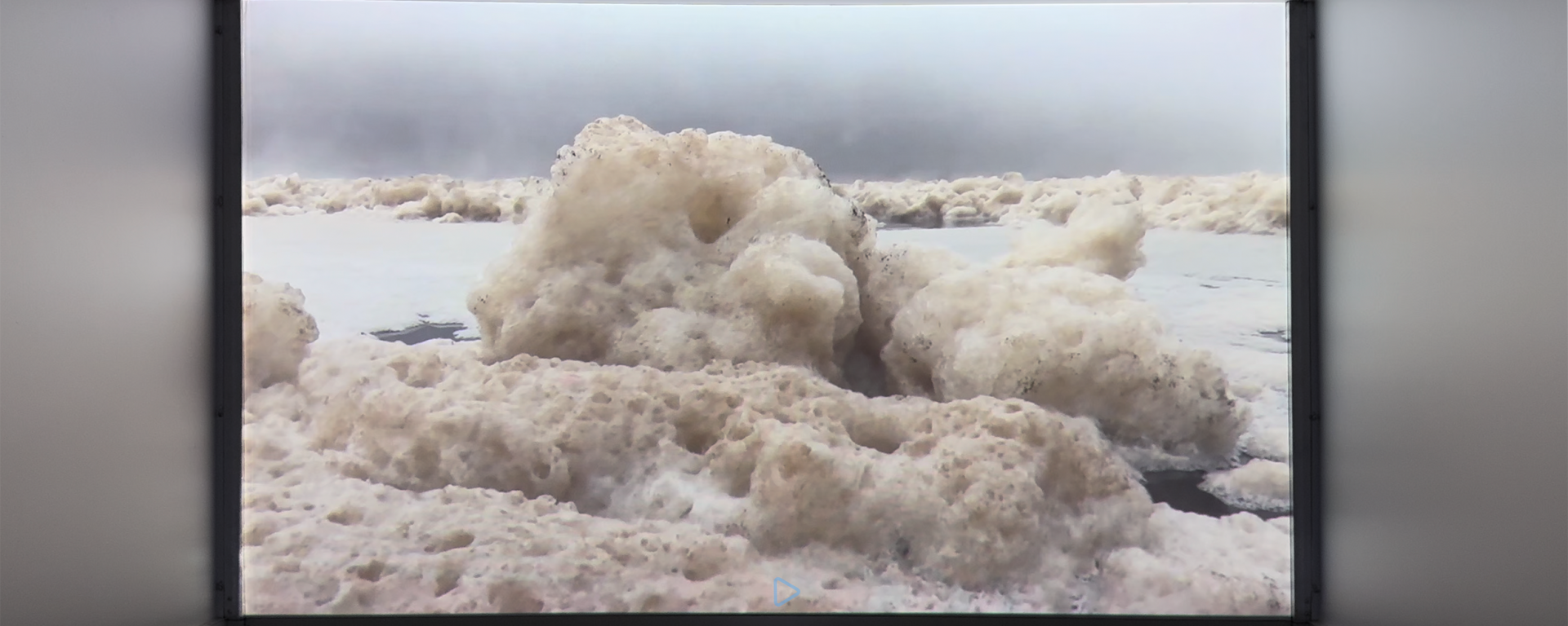 Aufnahme einer Videoprojektion auf Leinwand in einem Ausstellungsraum. Das Video zeigt eine winterliche Wasserlandschaft: Große Eis- und Schneemassen treiben auf einer vereisten Wasserfläche unter einem grauen, nebligen Himmel.