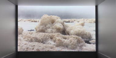 A video projection on a screen in an exhibition space. The video shows a wintry waterscape: large masses of ice and snow drift across a frozen body of water beneath a gray, foggy sky.