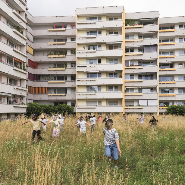 Kinder mit Tiermasken spielen auf einer Wiese vor einem Wohnblock.