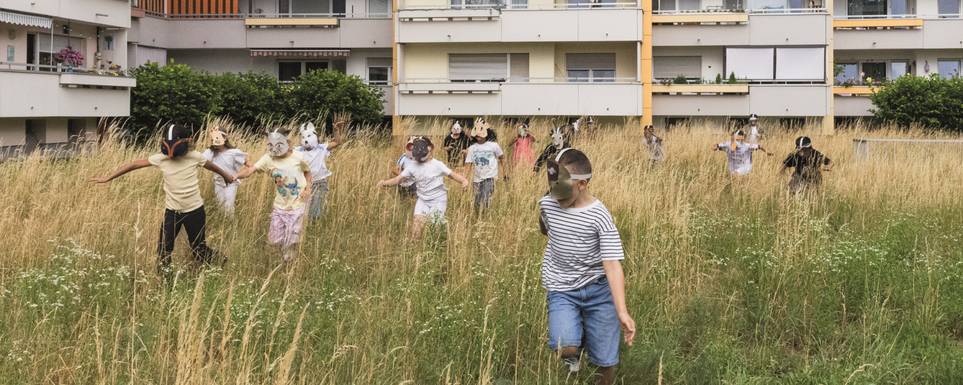 Kinder mit Tiermasken spielen auf einer Wiese vor einem Wohnblock.