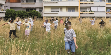 Kinder mit Tiermasken spielen auf einer Wiese vor einem Wohnblock.
