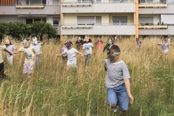 Kinder mit Tiermasken spielen auf einer Wiese vor einem Wohnblock.