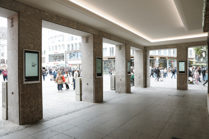 Überdachter Durchgang mit steinernen Säulen, Displays an den Säulen und Blick auf eine belebte Fußgängerzone.