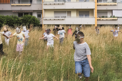 Kinder mit Tiermasken spielen auf einer Wiese vor einem Wohnblock.